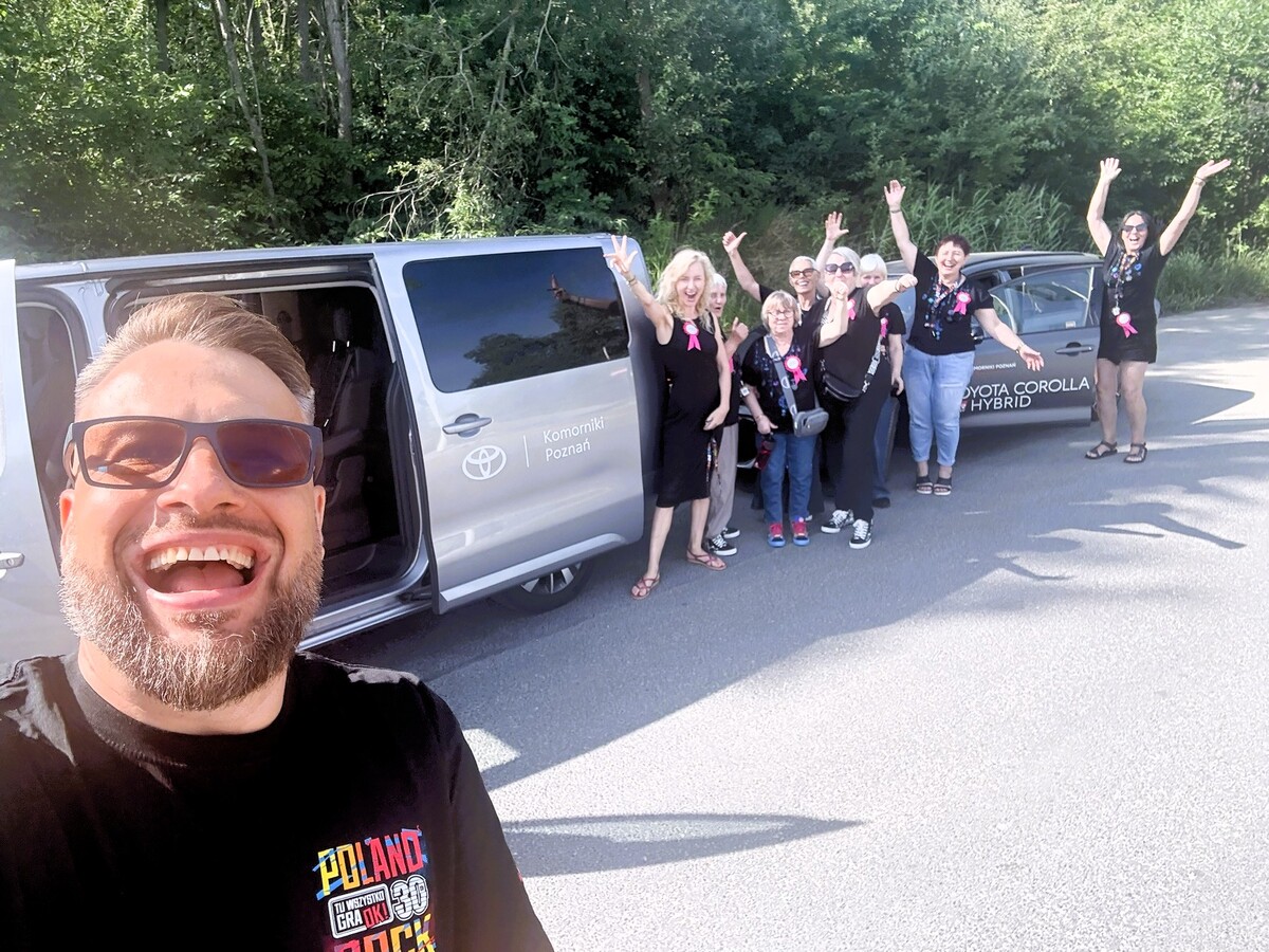 Wolontariusz robi radosne selfie przy dwóch samochodach Toyoty. Za nim grupa Mam z różowymi przypinkami „Oddech dla MAM” macha do aparatu i unosi ręce przed dalszą podróżą.